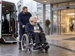 Transportmitarbeiter begleitet eine ältere Frau im Rollstuhl vor einem Klinikeingang; daneben steht ein Transportfahrzeug.